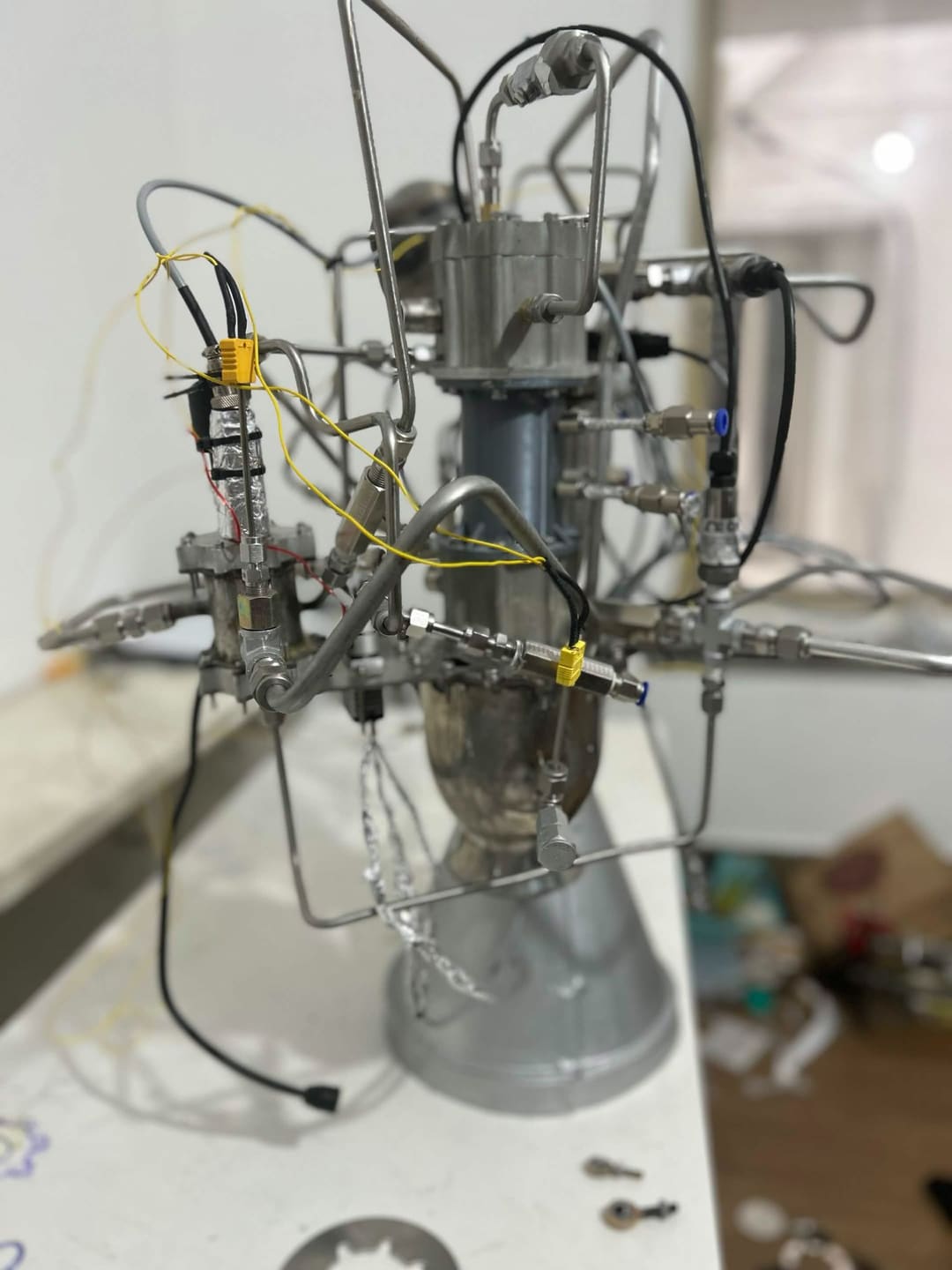 Front view of APEX propulsion development hardware on a bench, with tubing, cabling and yellow connectors against a blurred background.